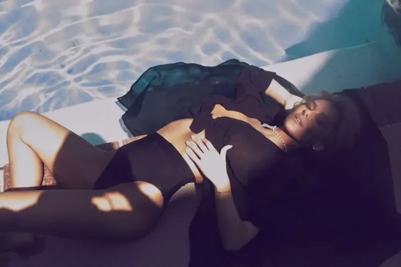 Katherine Taylor reclining on a pool lounge in a black swimsuit and pearls