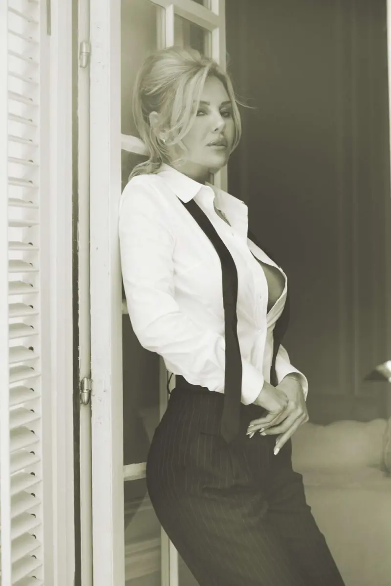 Black-and-white portrait of Katherine Taylor in a white shirt, suspenders, and pinstripe trousers beside open shutters