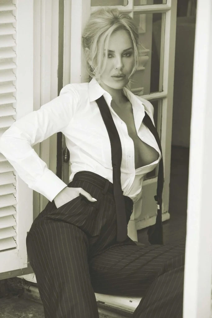 Black-and-white photograph of Katherine Taylor in an open white shirt, dark tie, and pinstripe trousers, seated in a doorway